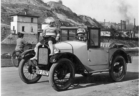 A black-and-white photograph of a small open tourer with its hood folded, a small child perched on the bonnet, a cyclist beside it.