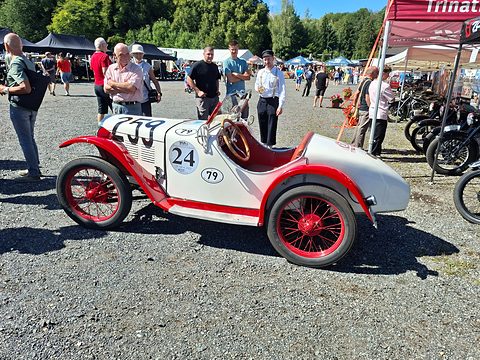 Side view of a white two-seater with red wings and wheels and racing numbers, visitors on the gravel around it. Opens a larger view