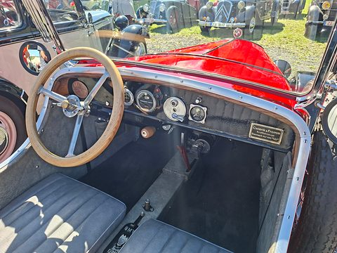 A cream steering wheel before a dashboard with round gauges and a small brass plaque, grey seats below. Opens a larger view