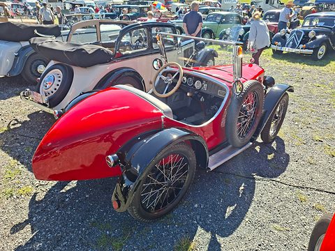 A red roadster with a long pointed tail, black wings and a spare wheel at its side, parked on gravel. Opens a larger view