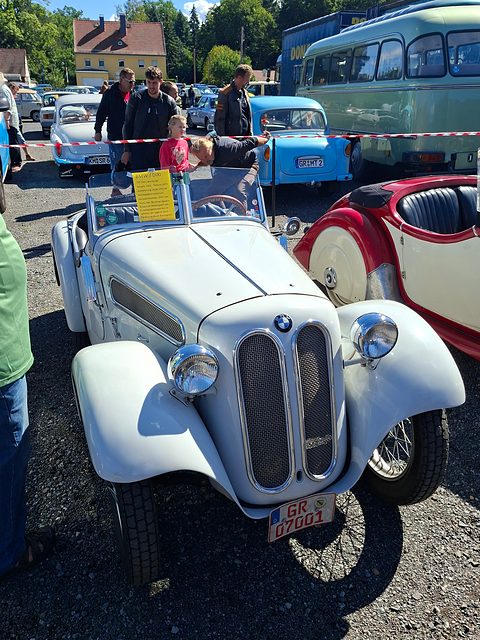 A white roadster with a split oval grille and wire wheels on gravel, visitors and a red car around it. Opens a larger view