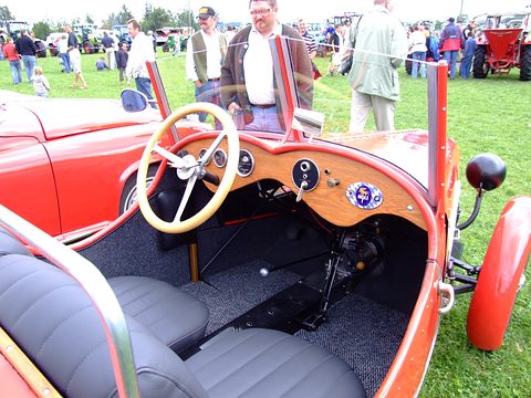 A cream steering wheel before a varnished wooden dashboard, grey seats and a gear lever, a crowd beyond. Opens a larger view