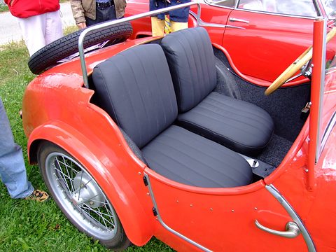 Two grey pleated seats in a low red open body with wire wheels, a spare wheel behind, on grass. Opens a larger view