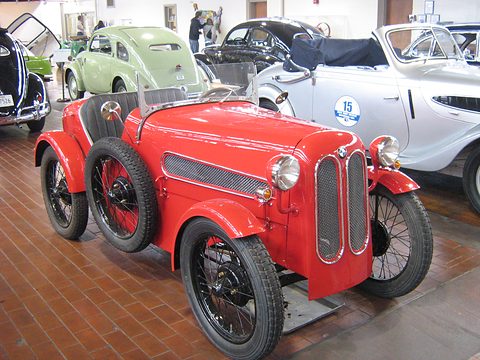 A red two-seat roadster with a split grille, louvred side and black wire wheels on a brick floor. Opens a larger view