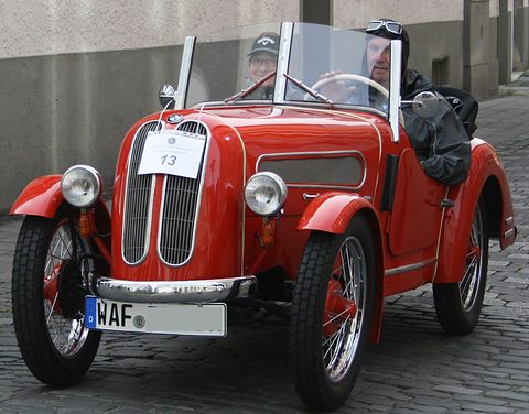 A red roadster with a split grille and chrome bumper on cobbles, two men in goggles aboard. Opens a larger view