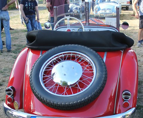 Rear view of a red roadster with a spare wire wheel and chrome hubcap set into its tail, a black hood folded. Opens a larger view