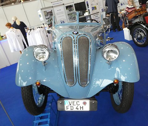 Front view of a pale blue roadster with a split oval mesh grille between two headlamps on a blue show carpet. Opens a larger view
