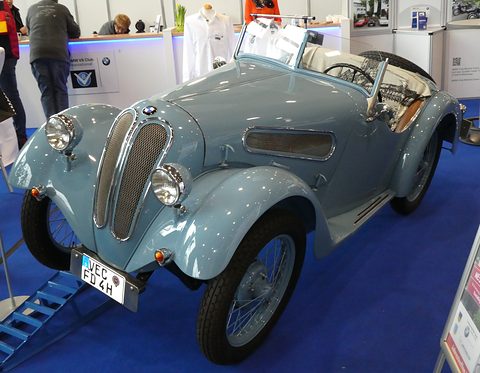A pale blue two-seat roadster with flowing wings, a split grille and wire wheels on a blue carpet. Opens a larger view