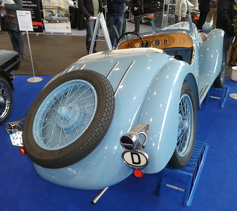 Rear view of a pale blue roadster with a spare wire wheel set into its rounded tail and a D plate. Opens a larger view