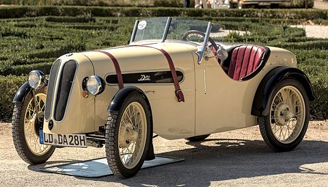 Side view of a cream sports two-seater with burgundy leather straps, red seats and cream wire wheels. Opens a larger view