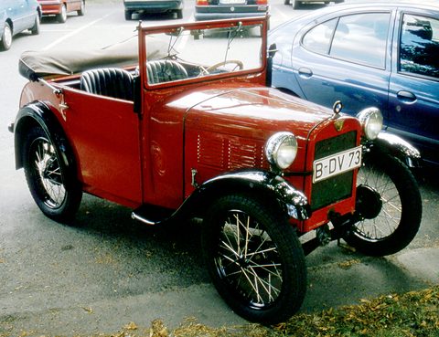 A red open tourer with a black radiator and wire wheels parked between modern cars in a car park. Opens a larger view
