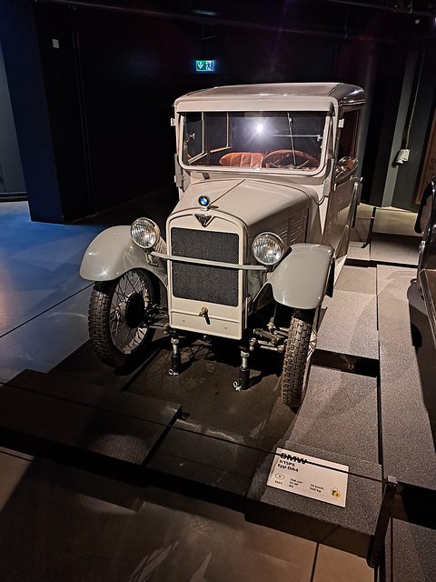 A grey-beige saloon with a chrome radiator spotlit on a dark stepped display, a label at its foot. Opens a larger view
