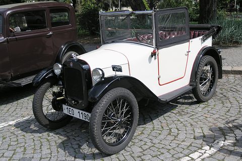 A white open tourer with black wings, a black radiator and wire wheels on cobbles beside a brown saloon. Opens a larger view