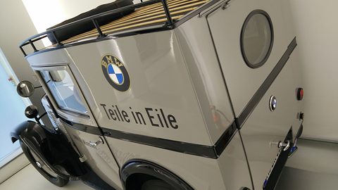 The grey side of a van with a roundel, the words Teile in Eile, a porthole window and a wooden roof rack. Opens a larger view