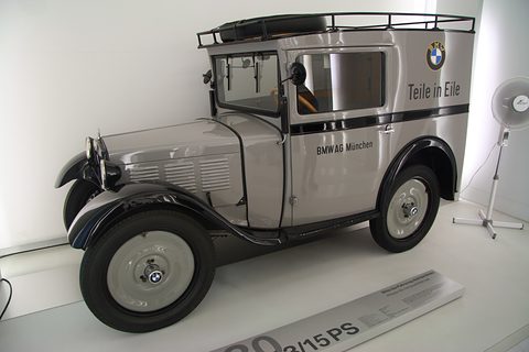 A small grey box van with a roof rack, black wings and BMW roundels on its sides in a white gallery. Opens a larger view