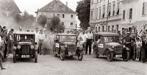 Three upright small saloons with numbered placards in their windscreens before a cheering crowd in a town square. Opens a larger view