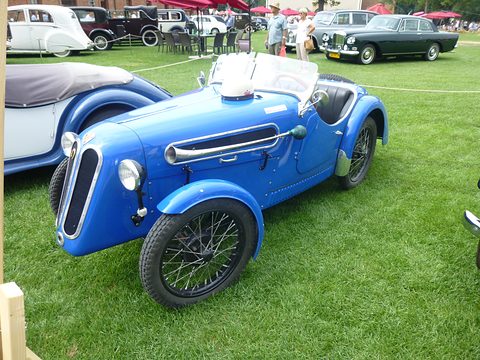 A bright blue roadster with a split oval grille, a louvred side panel and wire wheels on grass. Opens a larger view