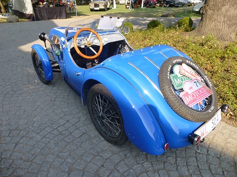 Rear three-quarter view of a blue roadster with a rally-plated spare wheel set into its rounded tail. Opens a larger view