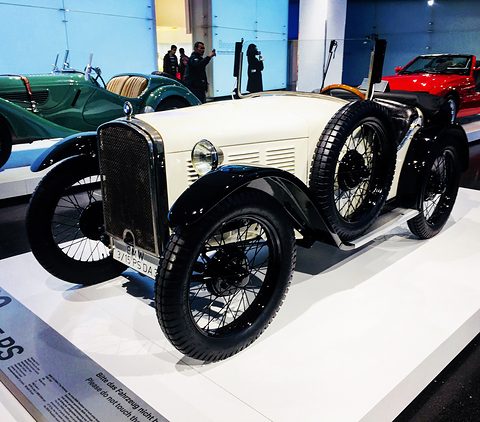 A white open two-seater with black wings and wire wheels on a white plinth with a lettered edge. Opens a larger view