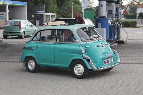 Turquoise bubble car pulling away across paving past fuel pumps, a driver visible through the side windows. Opens a larger view