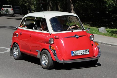 Red car with white roof turning on a tree-shaded road, two occupants visible, chrome bumper and front door handle. Opens a larger view