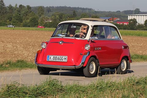 Red car with cream roof driving along a field road towards the camera, fabric roof rolled back, fields behind.