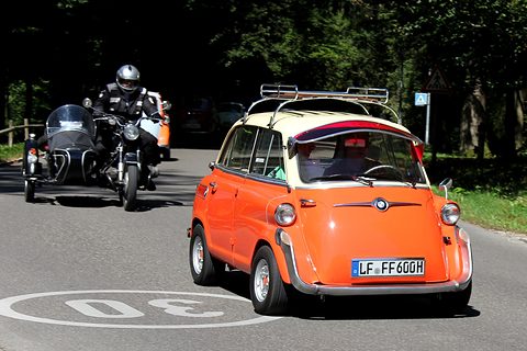 Orange car with cream roof and roof rack driving towards the camera past a 30 marking, a motorcycle and sidecar behind. Opens a larger view