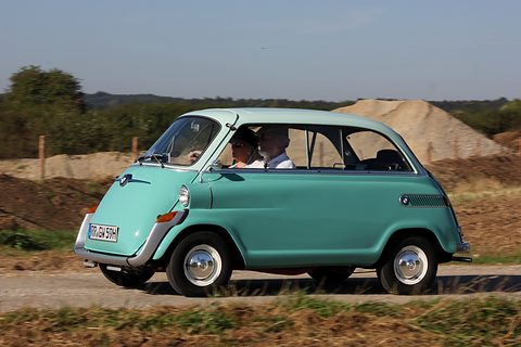 Turquoise small car with a pale green roof driving past a sand heap and fields, two occupants in sunglasses. Opens a larger view