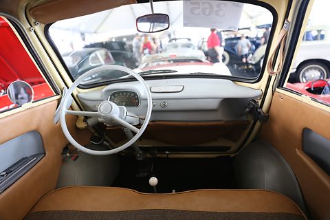 View into a small car through its open front: thin-rimmed white wheel, grey dash, gear lever and tan seat. Opens a larger view