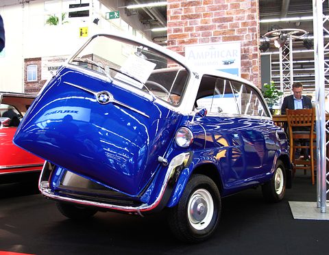 Glossy blue bubble car with white roof, its front door tilted open, on black carpet at a motor show stand. Opens a larger view