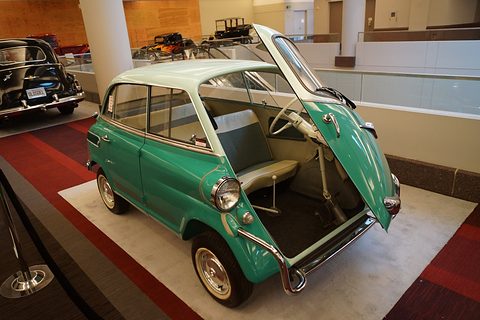 Green small car with pale roof on a carpet, whole front panel hinged open showing the wheel, bench seat and gear lever. Opens a larger view
