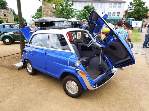 Blue bubble car with white roof, its front-hinged door swung up and the steering column coming with it, on gravel. Opens a larger view