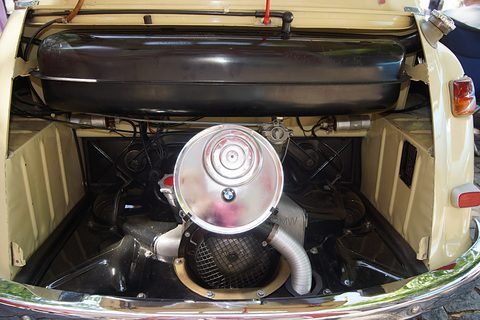 Open engine bay of a cream car: silver engine with BMW-badged air filter, mesh fan grille, black tank across the top. Opens a larger view