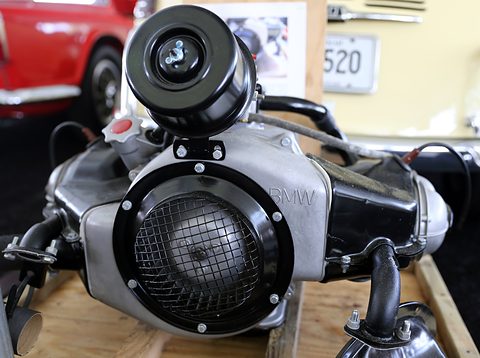 Silver and black two-cylinder engine on a wooden pallet, round mesh grille, air filter on top and BMW cast in the casing. Opens a larger view