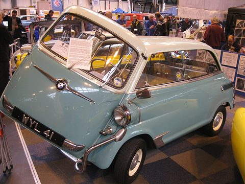Pale blue and cream small car at a crowded indoor show, roundel strip on the nose and a black number plate. Opens a larger view