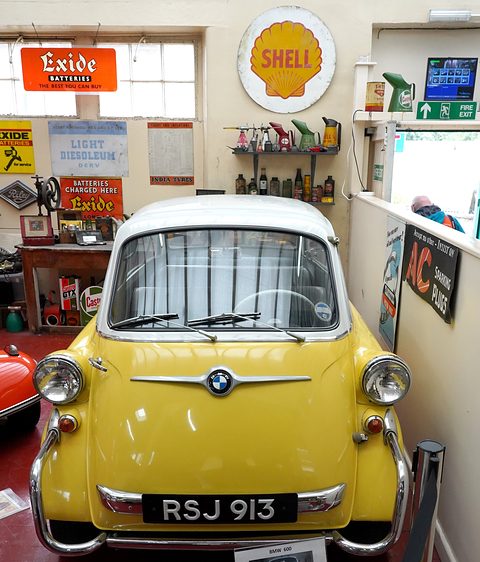 Head-on view of a yellow bubble car with white roof, black RSJ 913 plate, Shell and Exide signs on the wall behind. Opens a larger view