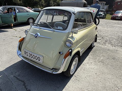 Pale cream bubble car with white roof on cracked tarmac in bright sun, other classic cars and people behind. Opens a larger view