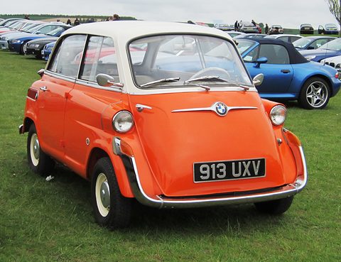 Orange bubble car with white roof parked on grass, rows of modern BMW roadsters behind under a grey sky. Opens a larger view