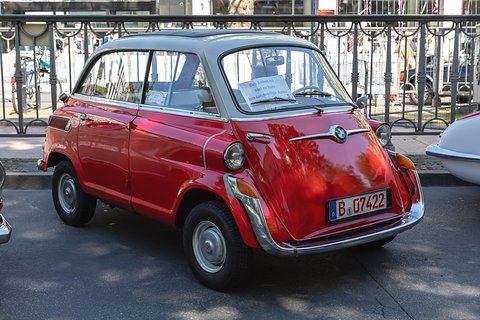Red small car with grey roof parked at a kerb, wrought-iron railings and spectators behind. Opens a larger view
