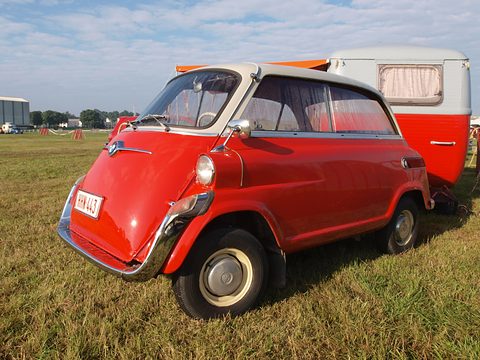 Glossy red small car with dew on its paint, parked on grass in front of a red and white caravan. Opens a larger view