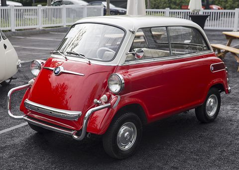 Red small car with white roof parked on dark tarmac, front door with roundel, picnic tables and a white fence behind. Opens a larger view
