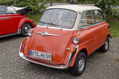 Orange bubble car with cream roof parked on cobbles beside a red Beetle cabriolet, hedges behind. Opens a larger view