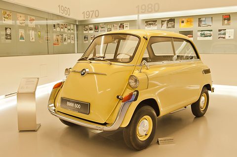Pale yellow four-wheeled bubble car with BMW roundel on its front door, on a museum floor before a wall of period adverts. Opens a larger view