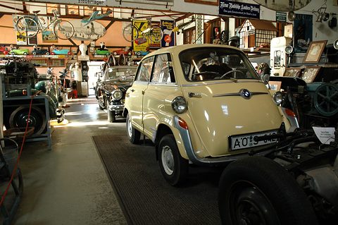 Cream bubble car with front door and BMW badge in a crowded hall of old cars, bicycles and enamel signs. Opens a larger view