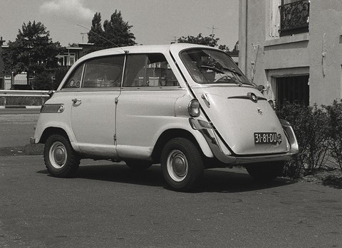 Black-and-white photograph of a pale bubble car parked by a house, side door hinges and front panel visible.
