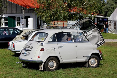 Off-white small car on grass seen from the rear right, nose panel raised, roof rack with a tan suitcase. Opens a larger view