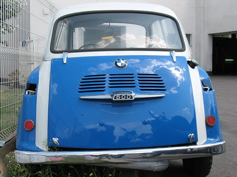Rear of a blue car with a white stripe on each side, louvred panel, round red reflectors and clouds reflected in the paint. Opens a larger view