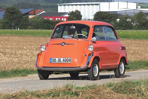 Orange bubble car coming straight towards the camera on a narrow road, stubble field and farm buildings behind. Opens a larger view