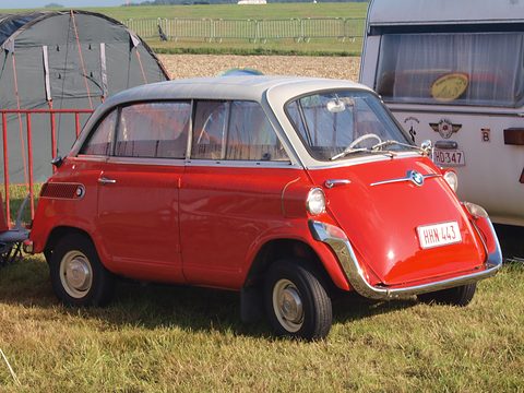 Red car with grey roof parked on grass between a dark green tent and a white caravan. Opens a larger view