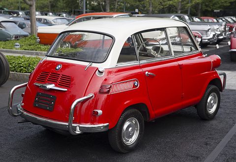 Rear three-quarter of a red car with white roof, chrome tubular bumpers, a rows of classic cars behind. Opens a larger view